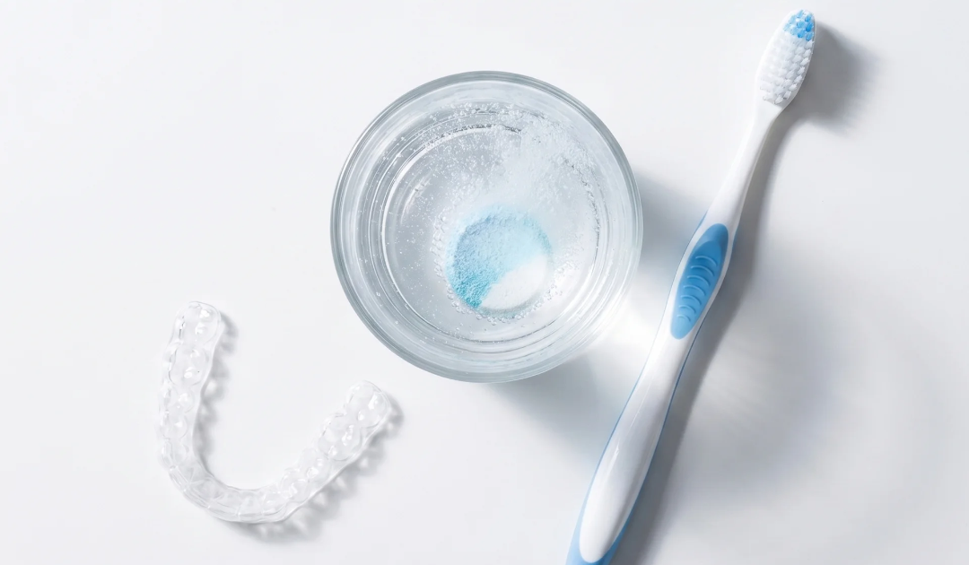 clear retainer, a small glass of water with a fizzing cleaning tablet dissolving in it, and a soft toothbrush, arranged neatly on a white surface