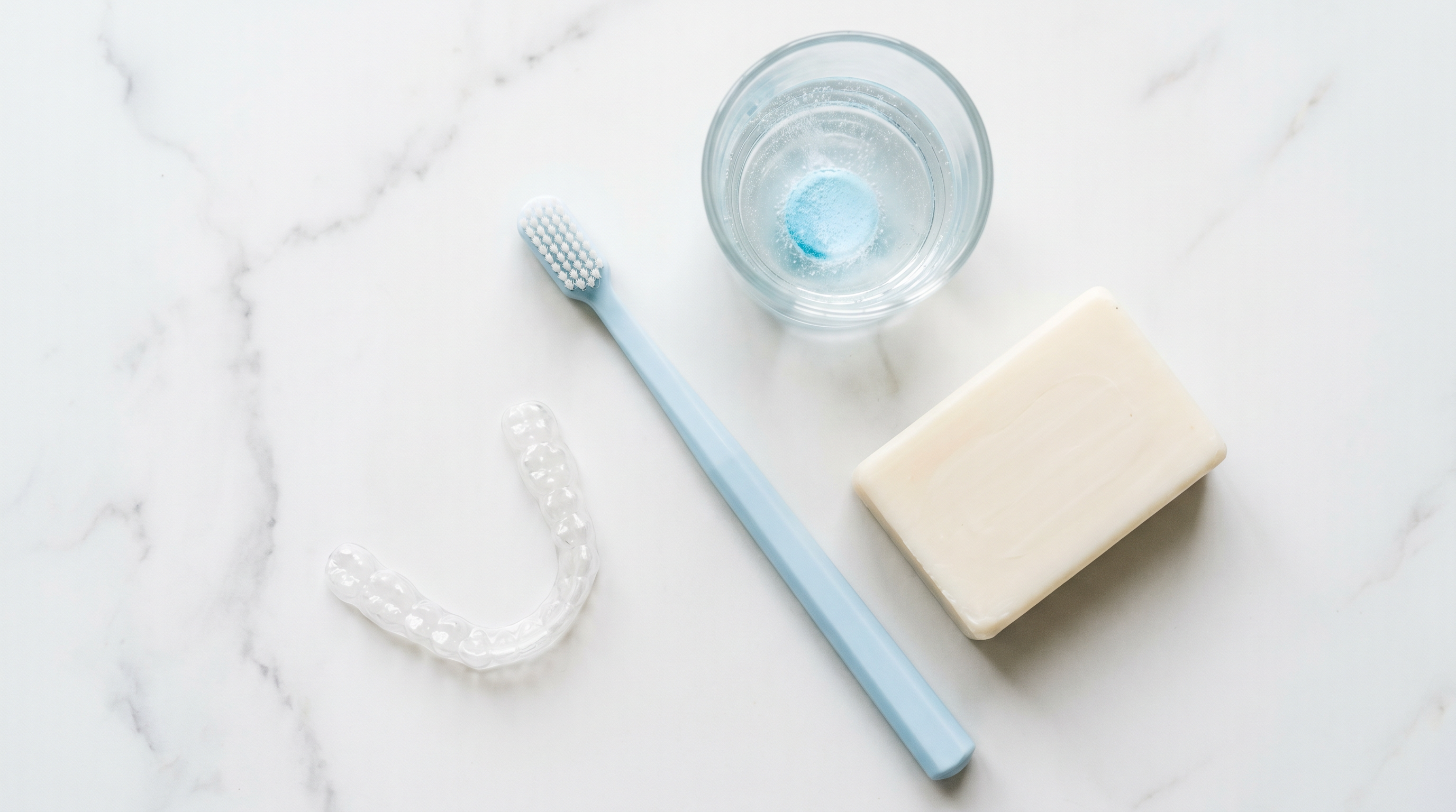 clear aligner tray toothbrush cleaning tablet and soap flat lay on white marble surface